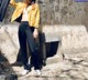 A woman in a yellow jacket and black pants leaning against a wall.