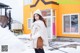 A woman standing in the snow in front of a yellow house.